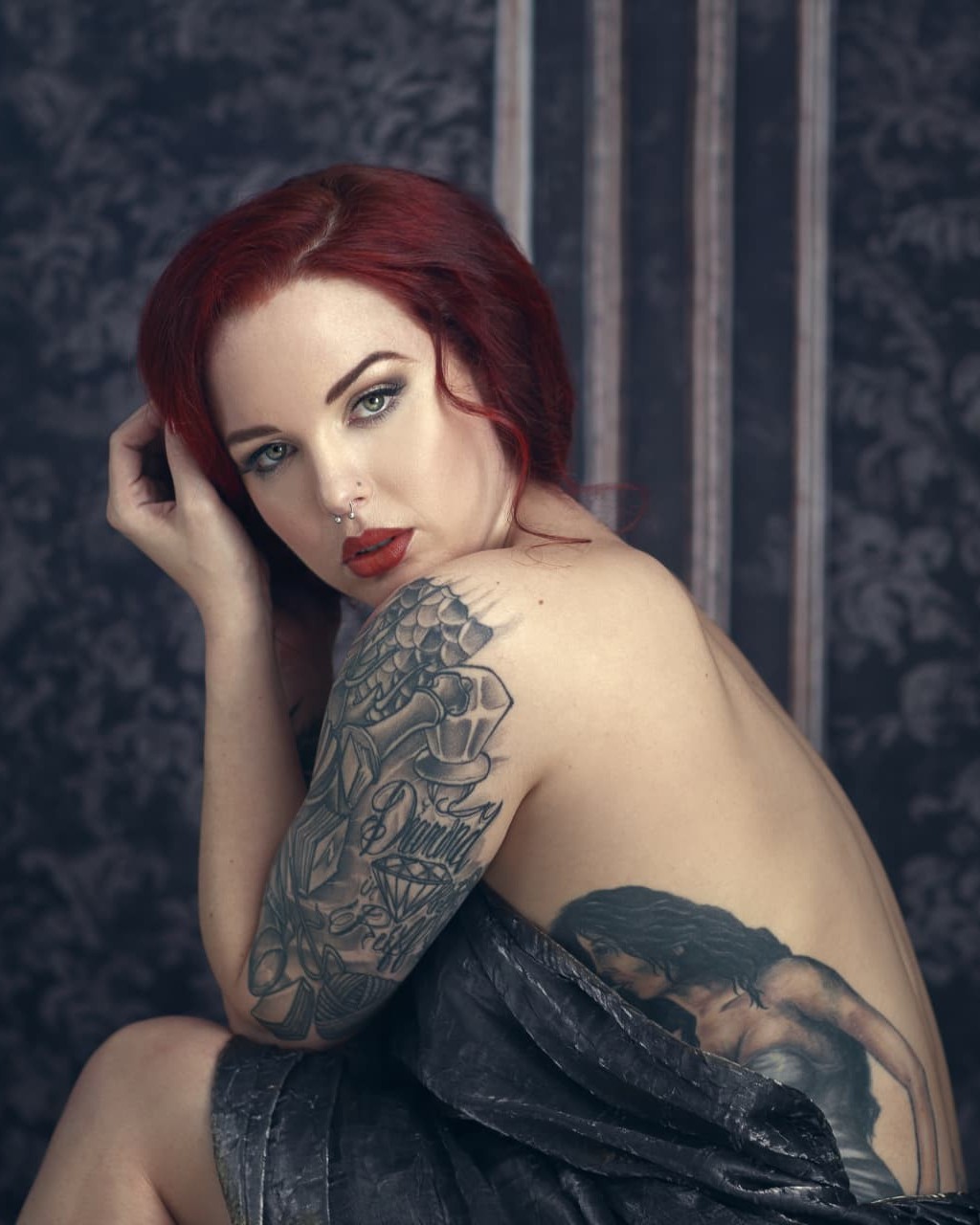 Boudoir client with red hair and tattoos looking back over her shoulder against a dark damask backdrop