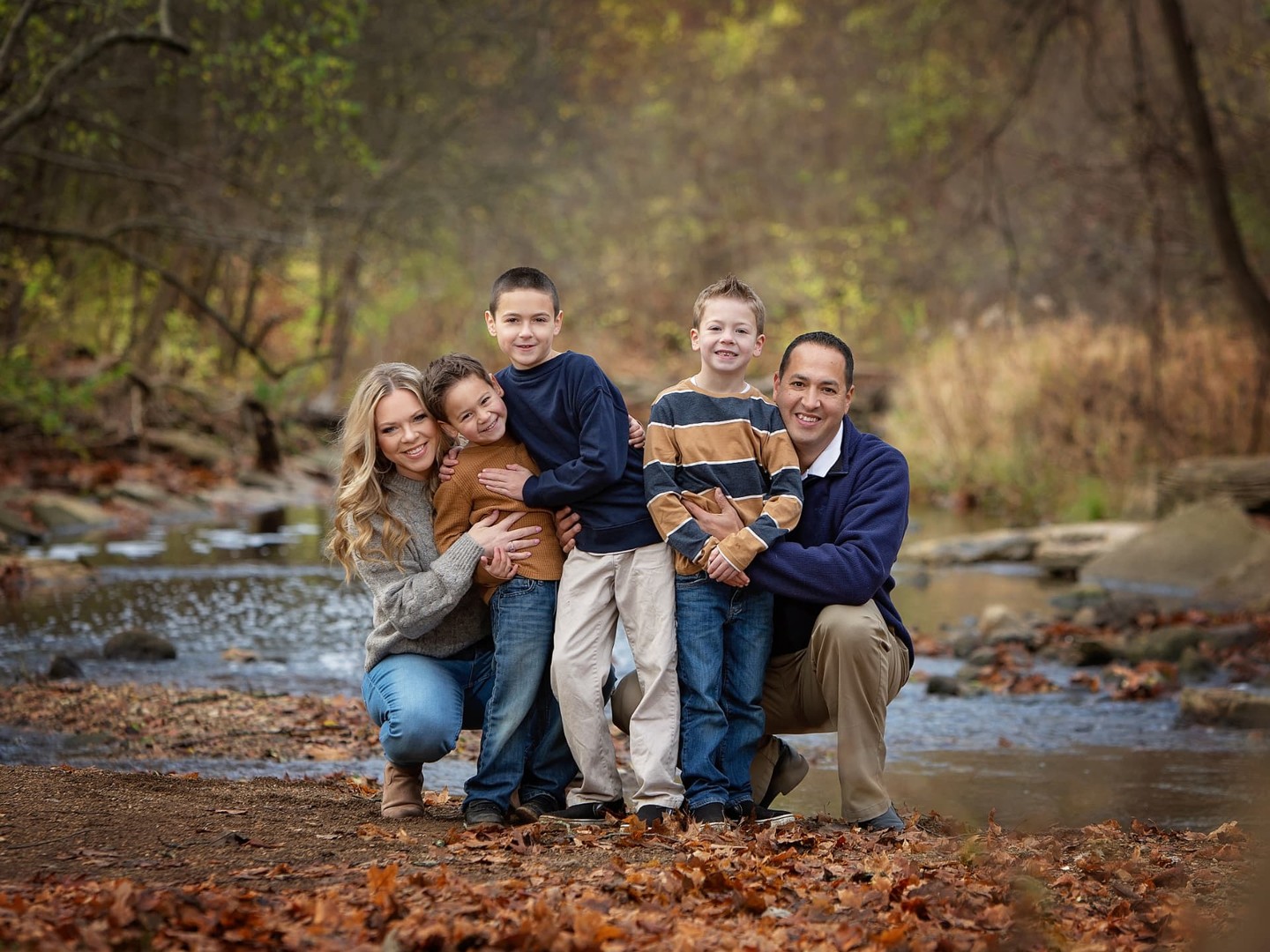 A mother, father, and their three boys posing together beside a creek in autumn woods