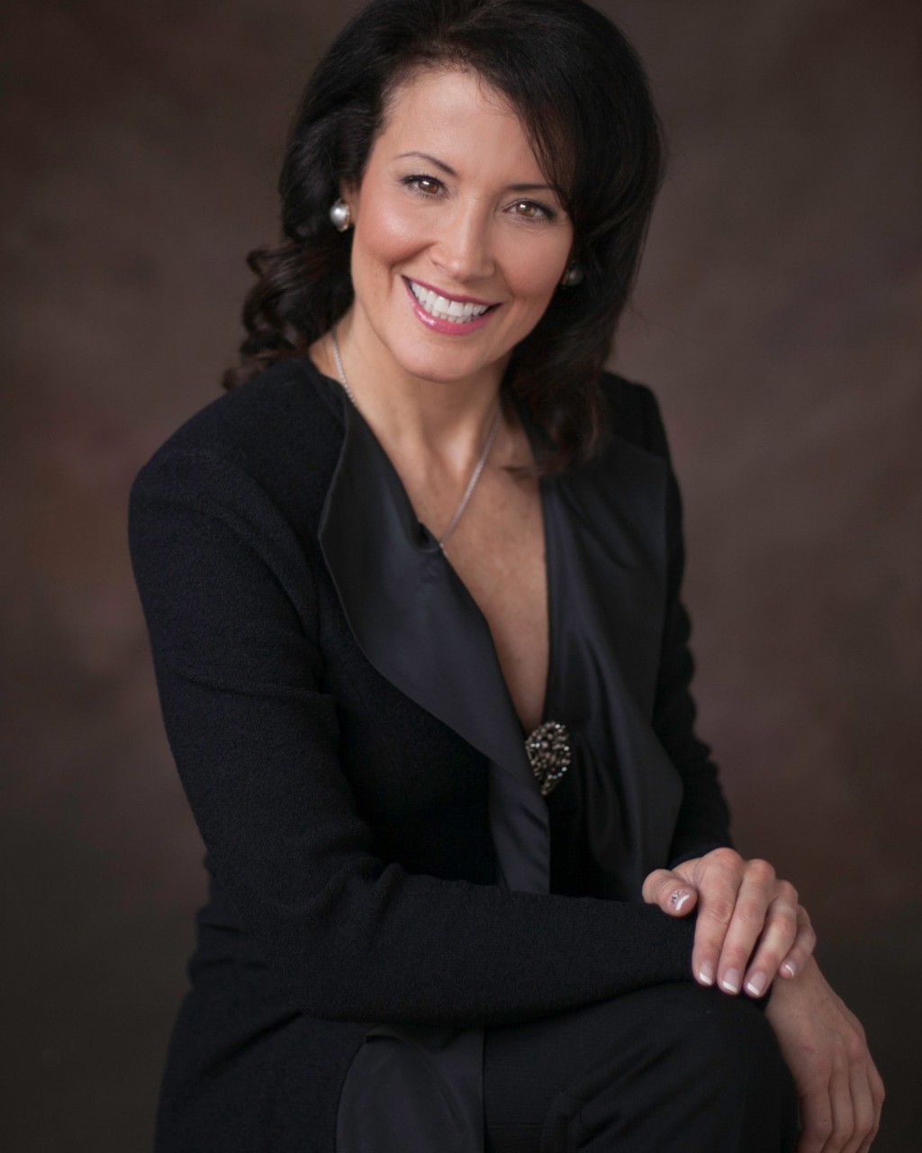 Professional headshot of a woman in a black jacket seated against a warm brown studio backdrop