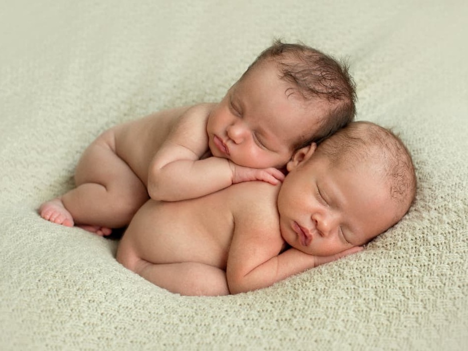 Two newborn twins sleeping together on a soft cream knit blanket