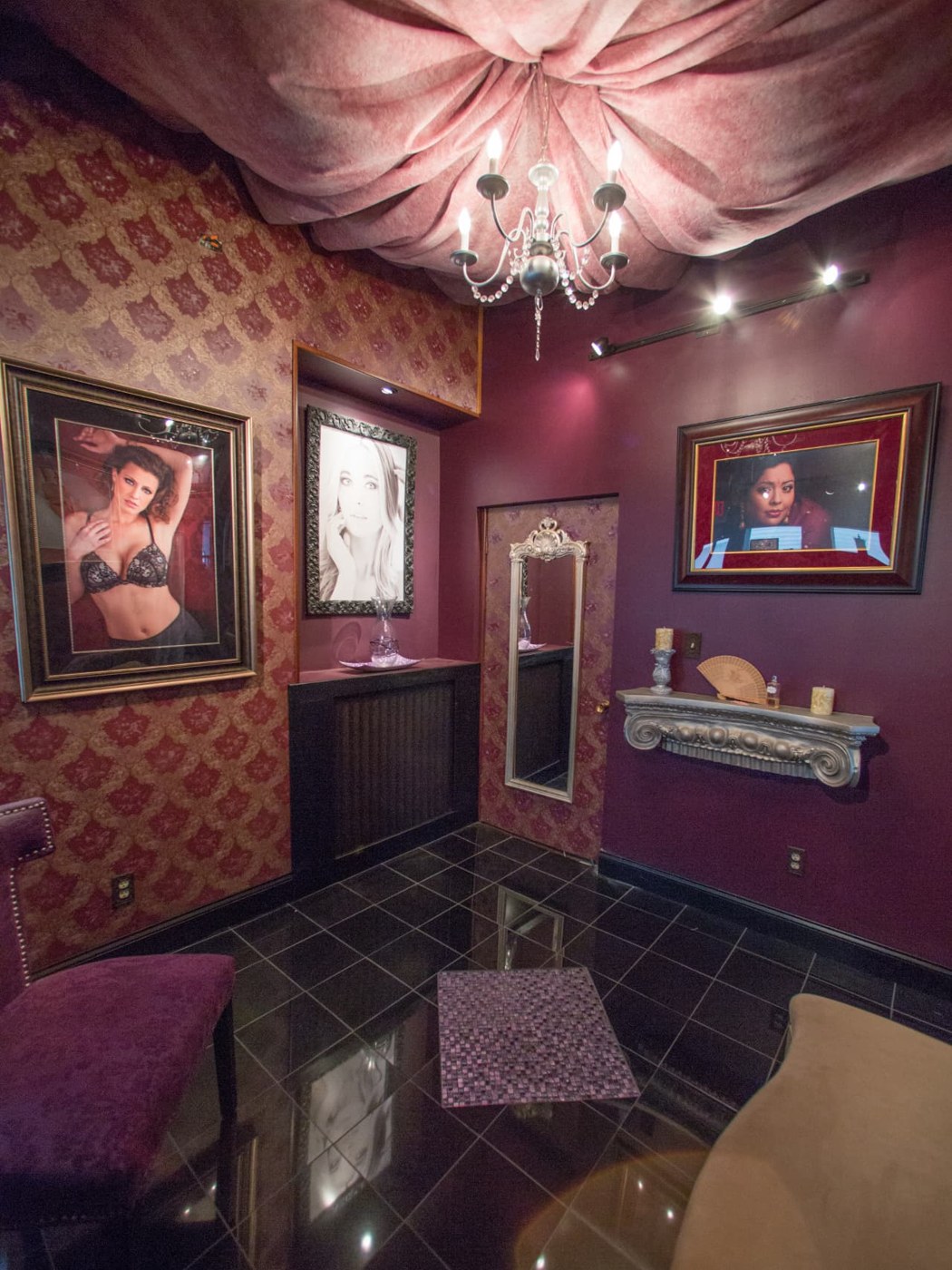 A damask-covered alcove with an ornate framed mirror, a carved wall shelf with candles, and framed portraits