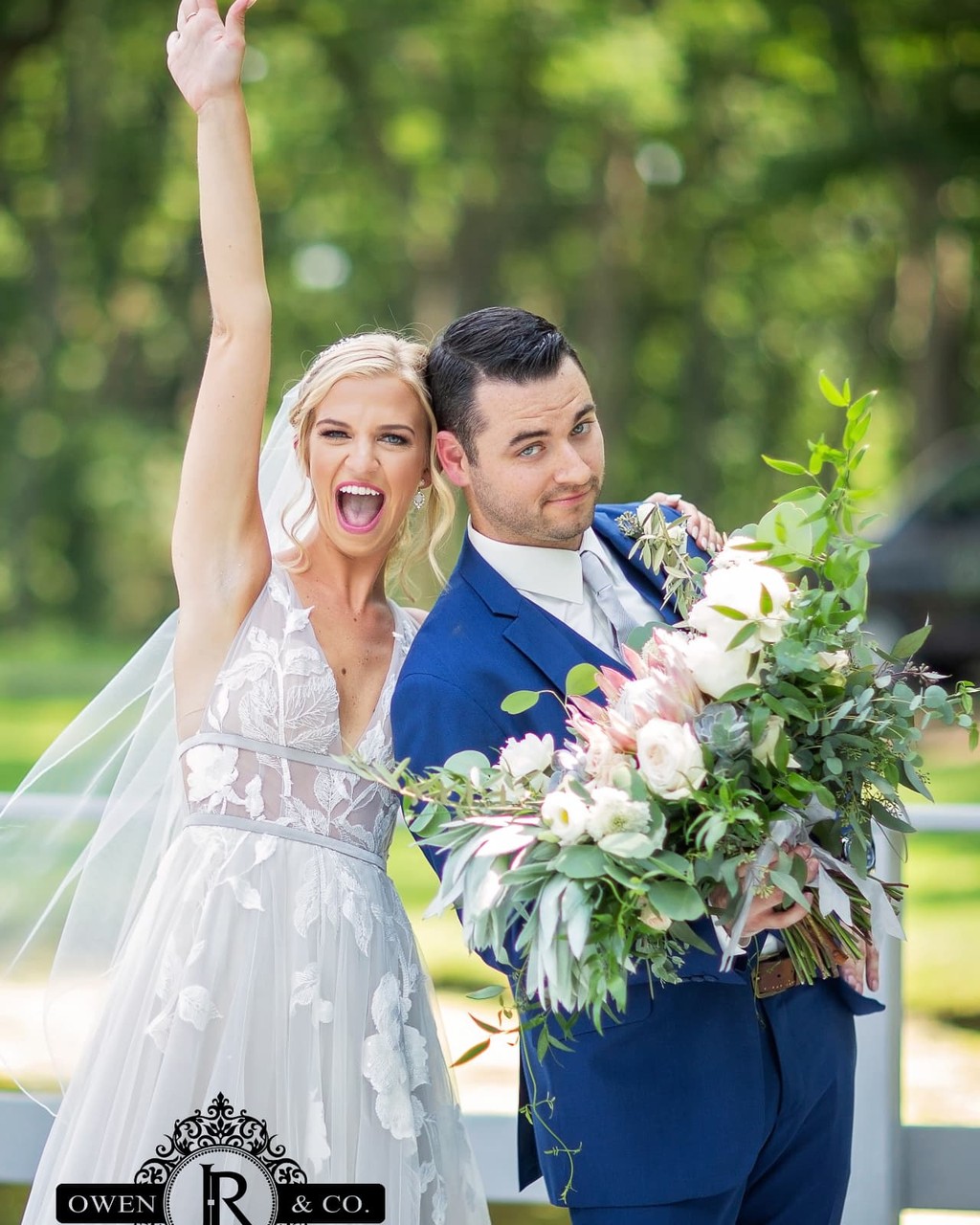 A bride with her arm raised in celebration beside a groom holding a large bouquet